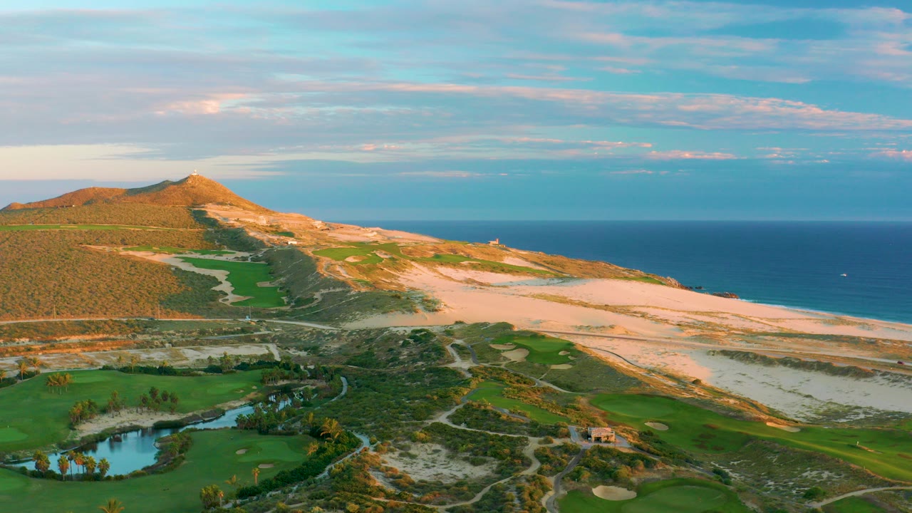 Wide aerial frame over Diamante Cabo San Lucas showing green golf fairways, a pond at lower left, sand dunes, a low hill, the ocean horizon, and no visible people.