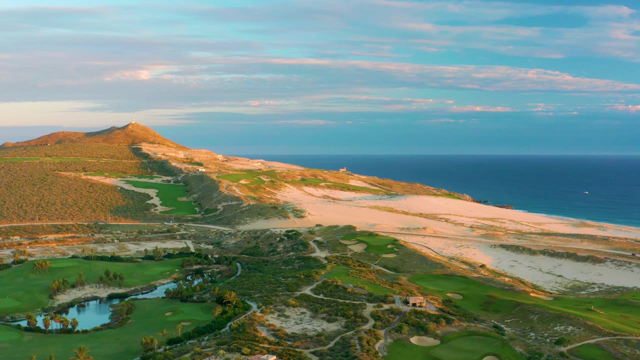 Wide drone-style frame of Diamante Cabo San Lucas with fairways and a pond below, sandy coastal terrain across the center, blue ocean to the right, low hills to the left, and no visible people.