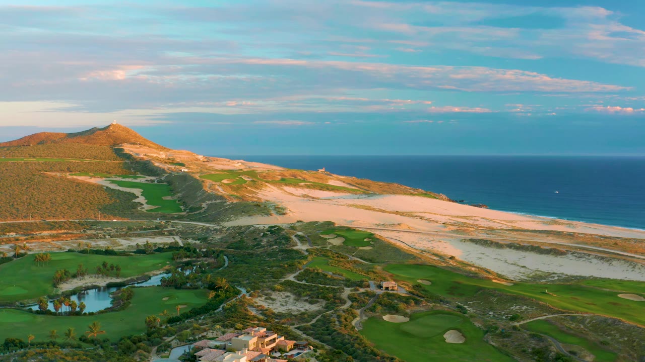 Aerial view at 0:01 over Diamante Cabo San Lucas with fairways and sand traps in the lower frame, pale dunes and beach across the center, and the ocean horizon behind, with no visible people.