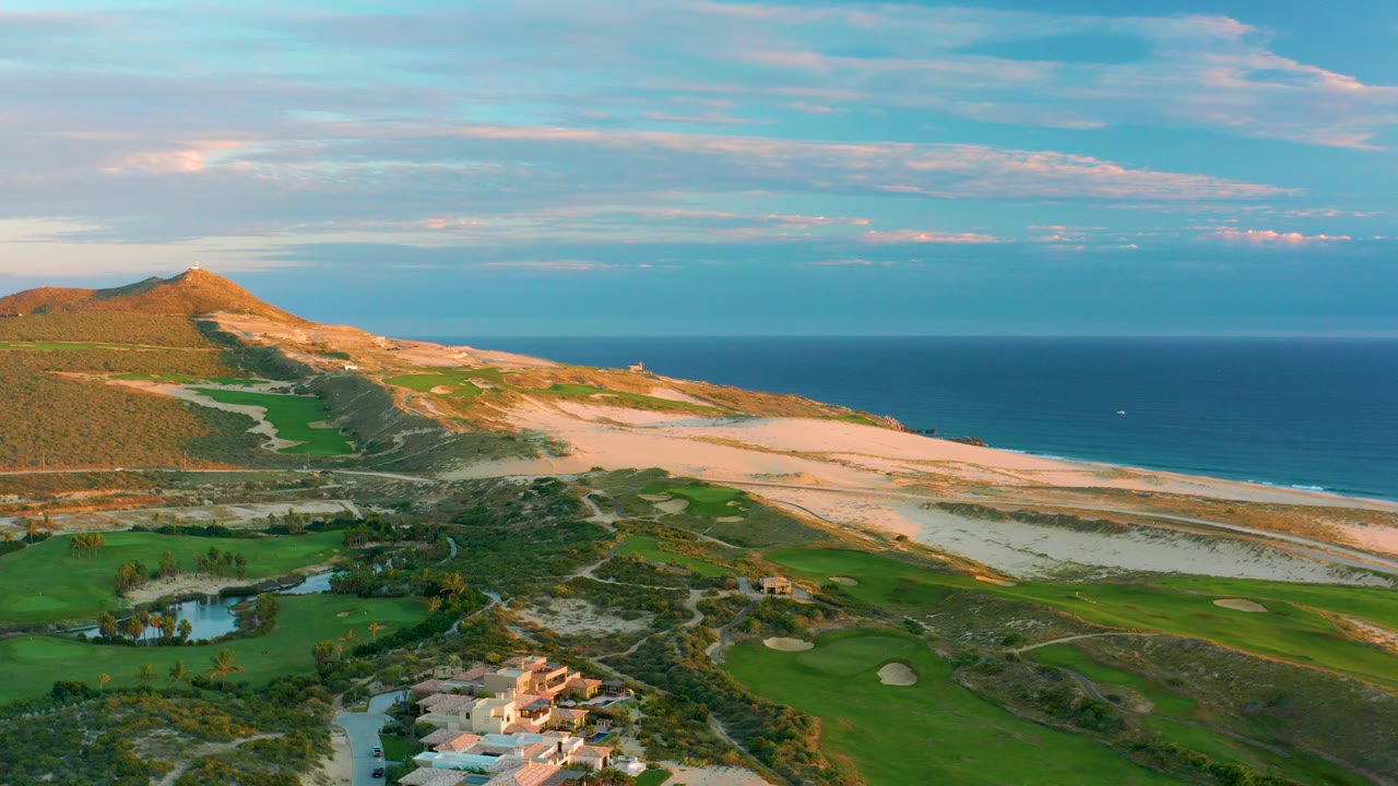 Aerial view over Diamante Cabo San Lucas with green golf fairways and water hazards in front, pale dunes and beach beyond, blue ocean on the horizon, and no visible people.