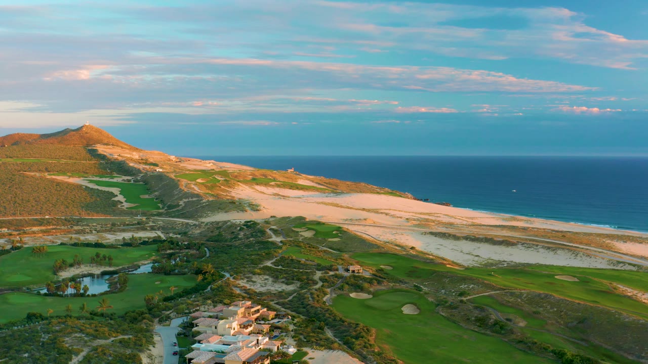 Wide aerial frame at Diamante Cabo San Lucas with sunlit sand dunes across the center, golf greens below, the calm ocean behind, and no visible people.