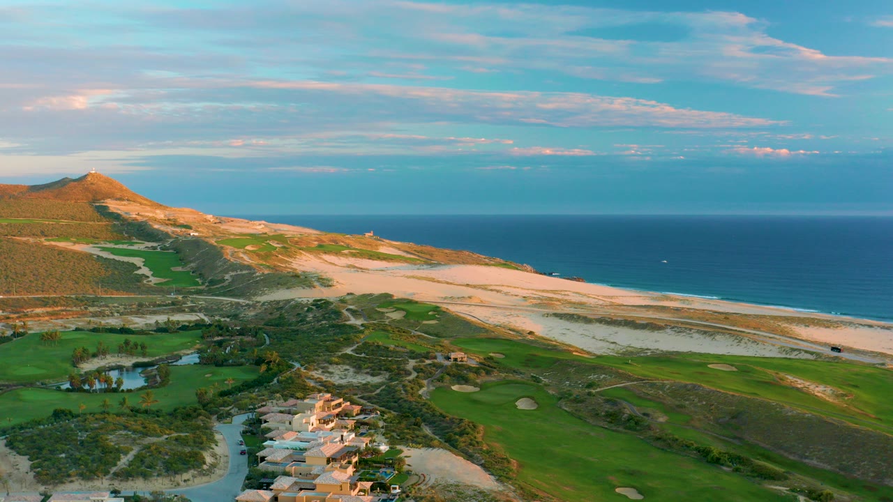 Aerial frame from Diamante Cabo San Lucas showing green golf fairways, sand traps, clustered coastal buildings at the lower edge, pale beach, blue ocean, and no visible people.