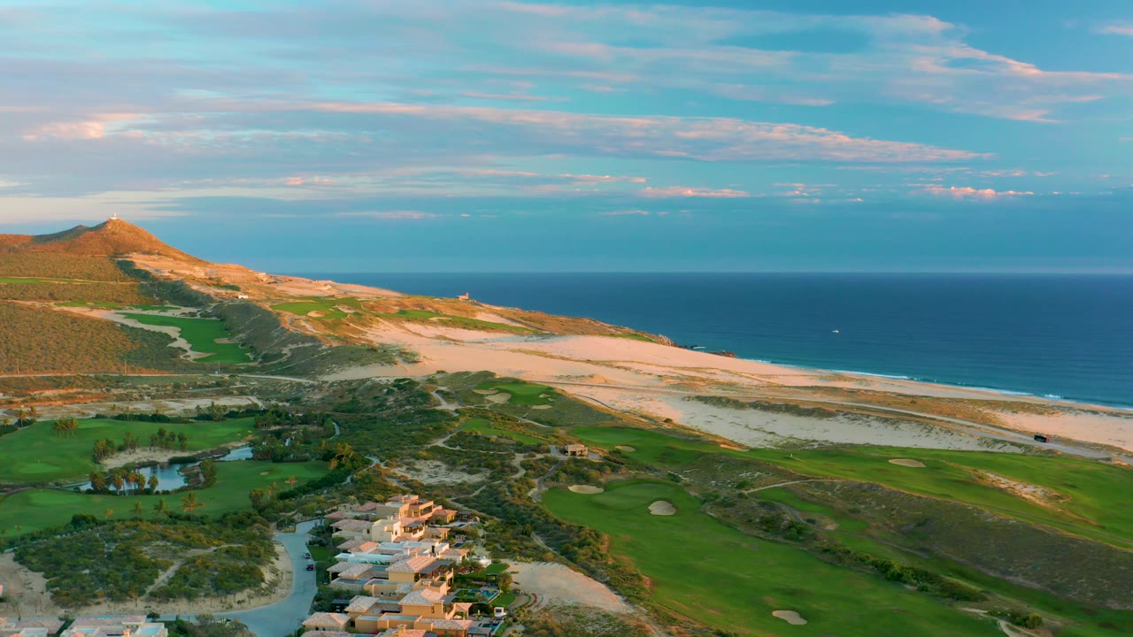 The aerial view over Diamante Cabo San Lucas keeps the hillside at left, golf greens below, pale beach through the middle, blue ocean beyond, and no visible people.