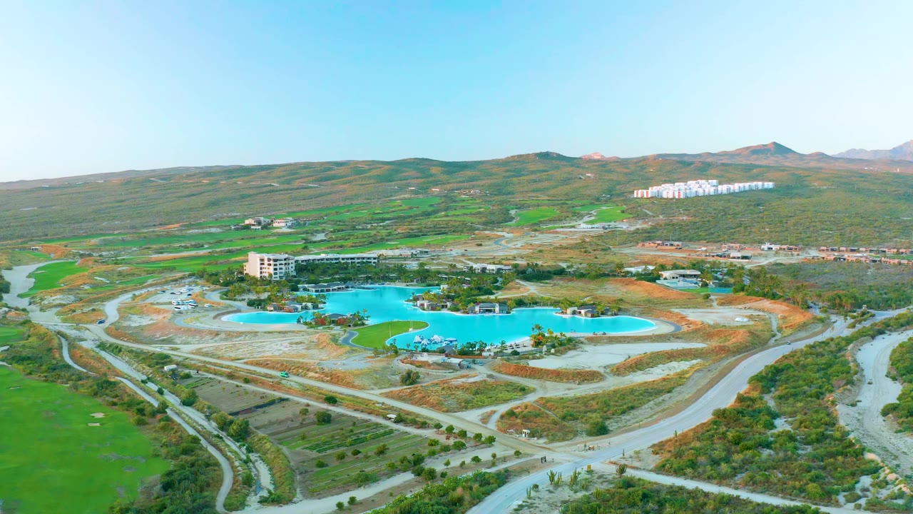 Aerial frame over Diamante Cabo San Lucas with the reservoir centered below the hillside, pale roads in the foreground, green fields to the left, and no visible people.