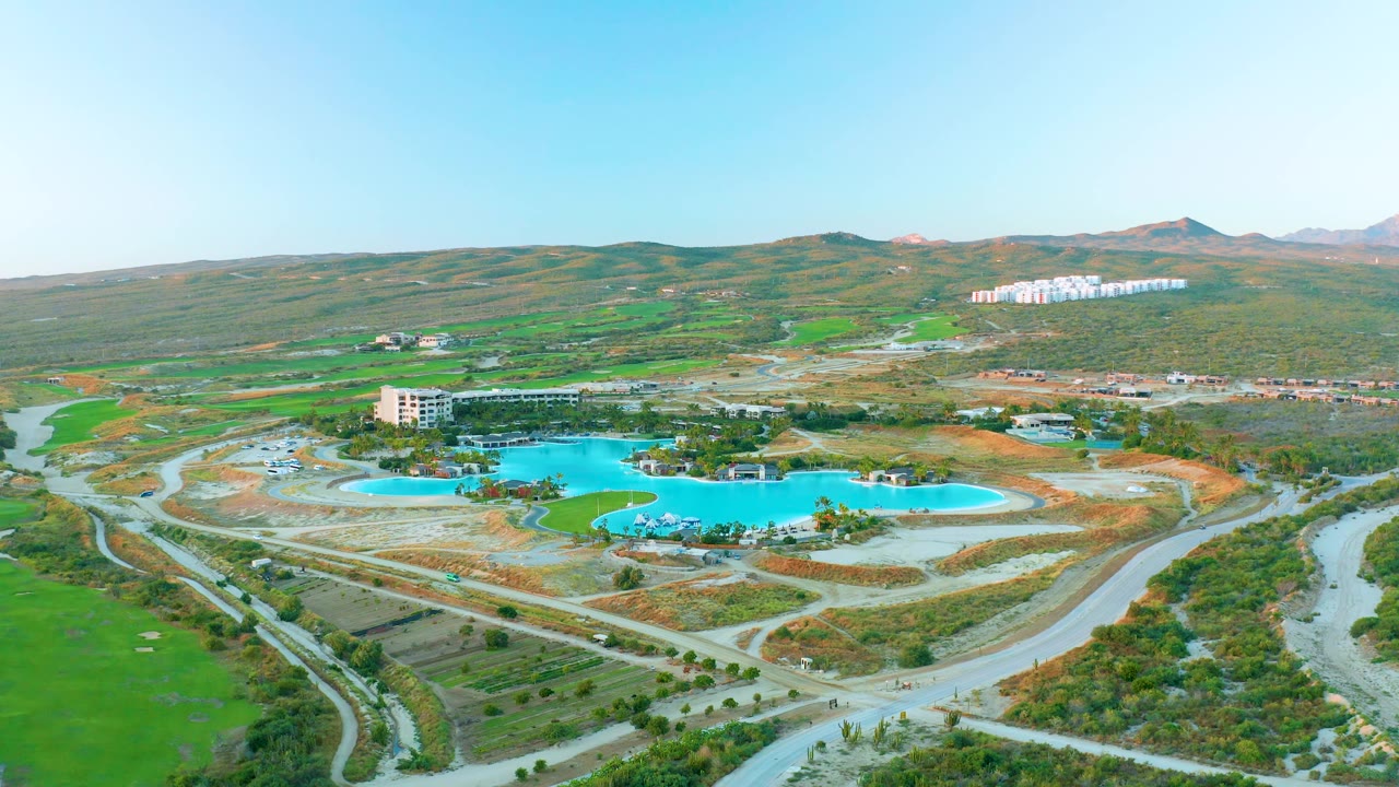 Drone frame in Diamante Cabo San Lucas showing blue reservoir water, nearby buildings, winding roads, green ground cover, low hills, and no visible people.
