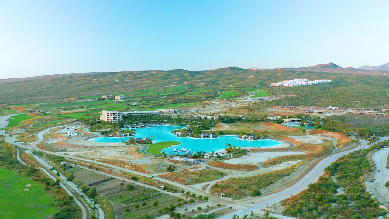 Early frame of Diamante Cabo San Lucas showing bright blue reservoir pools, curved construction roads, a low building, green hills, distant white structures, and no visible people.