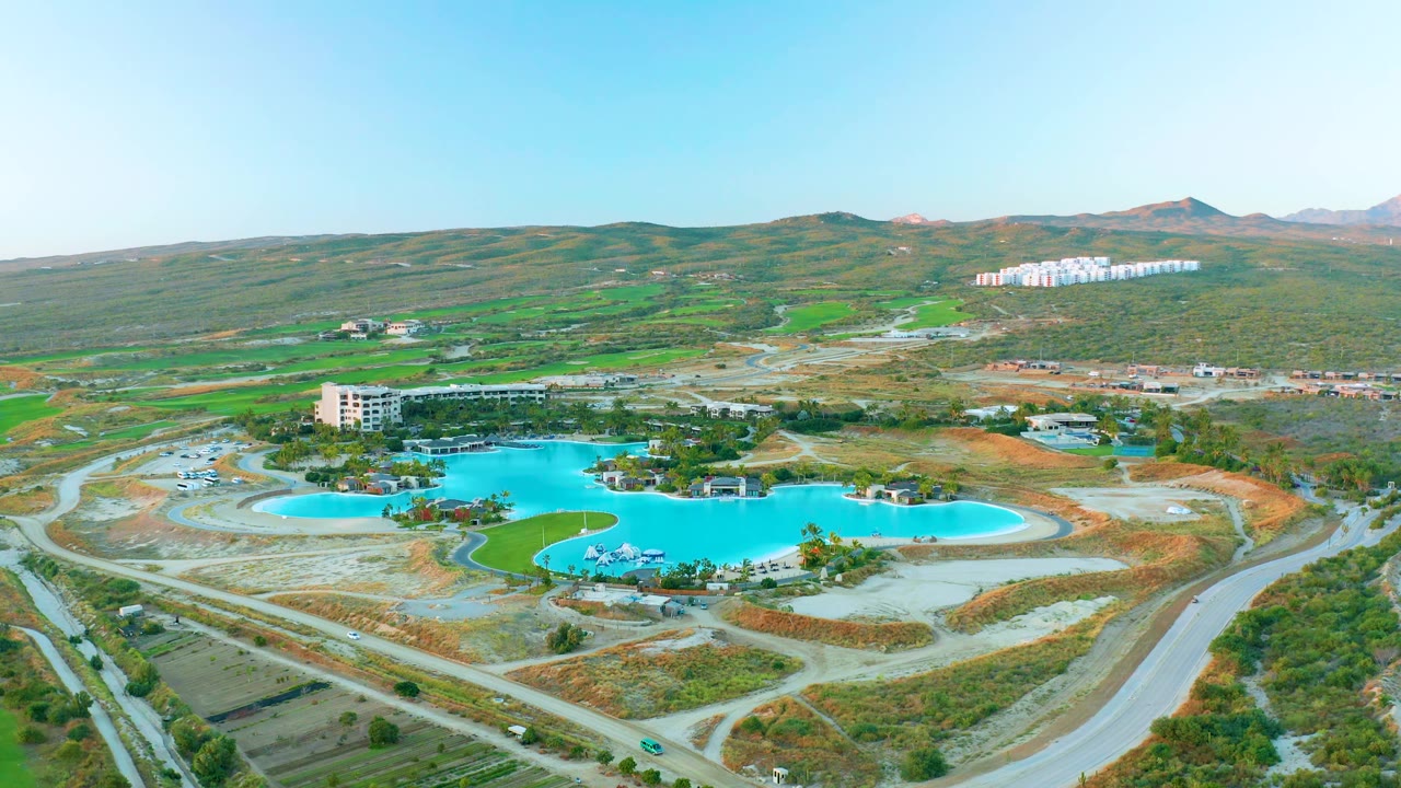 Drone view in Diamante Cabo San Lucas showing the reservoir centered below green hills, with pale road cuts, open lots, distant white housing blocks, and no visible people.