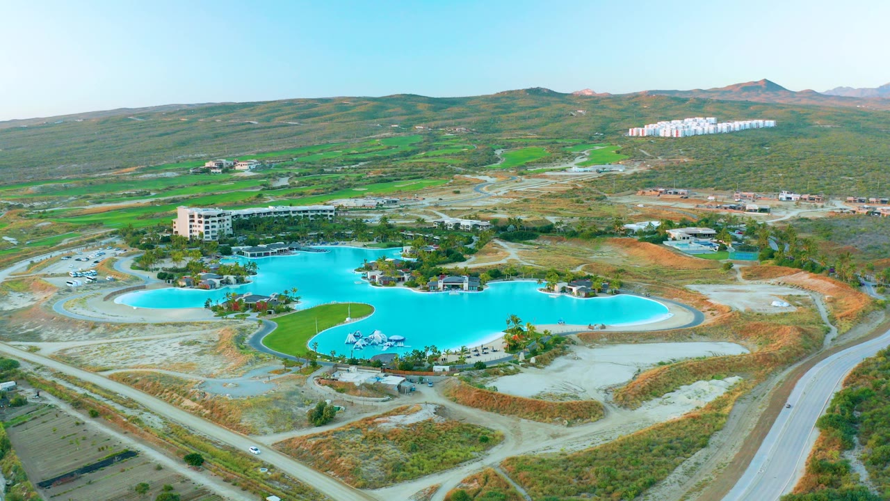 Early aerial frame over Diamante Cabo San Lucas showing a curved turquoise pool, pale roads, dry terrain, green patches, distant hillside buildings, and no visible people.