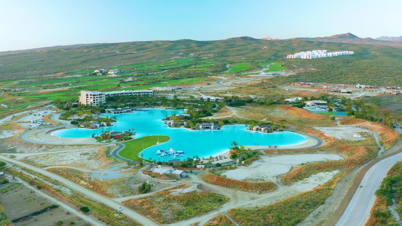 Mid-clip aerial view of Diamante Cabo San Lucas with turquoise pool water, circular road curves, low buildings, dry construction-like terrain, green patches on hills, and no visible people.