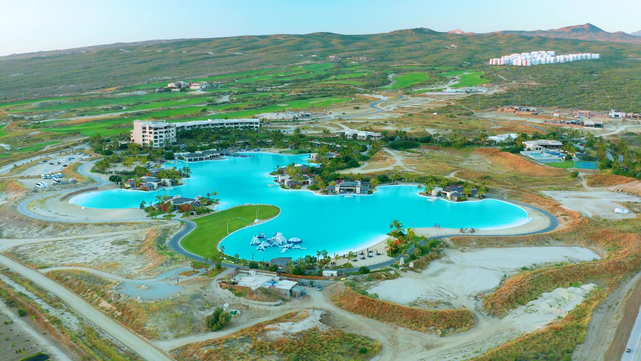 Opening aerial frame over a bright blue pool complex in Diamante Cabo San Lucas, with roads, exposed earth, green hills, low buildings, and no visible people.