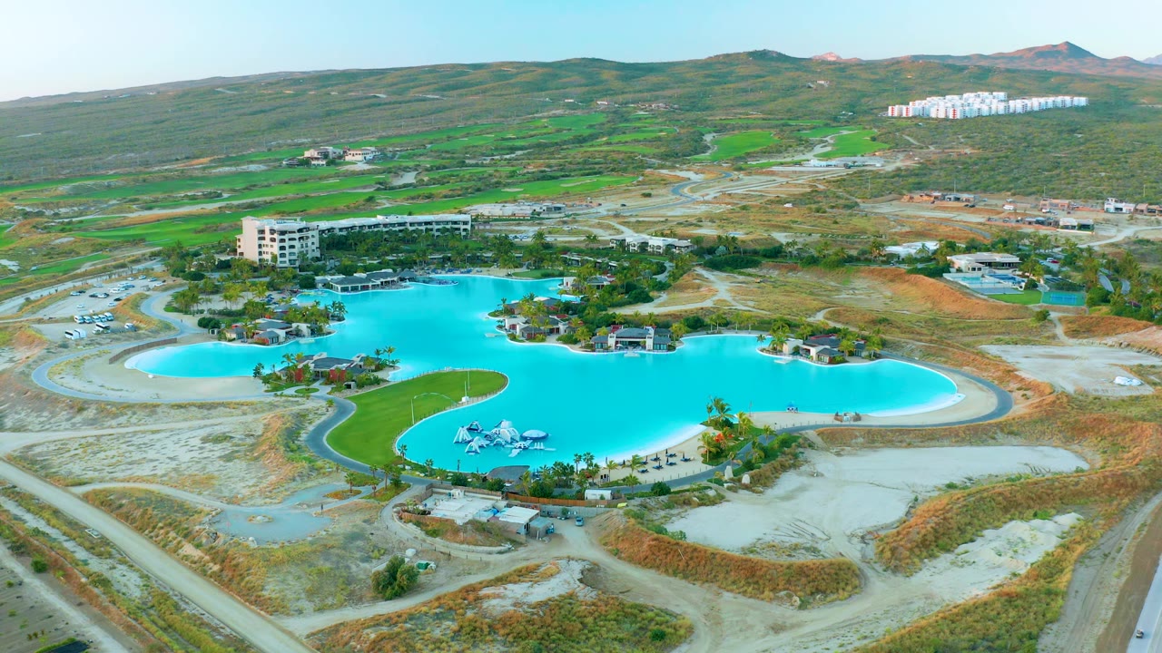 Second aerial frame of the Diamante Cabo San Lucas pool area, with turquoise water, curved road edges, landscaped grass, dry earthworks, hills, and no visible people.