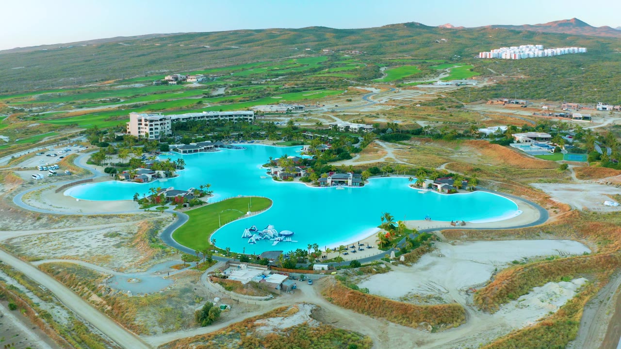 Third frame from above the pool complex in Diamante Cabo San Lucas, showing blue water, small poolside structures, a green lawn strip, hills, roads, and no visible people.