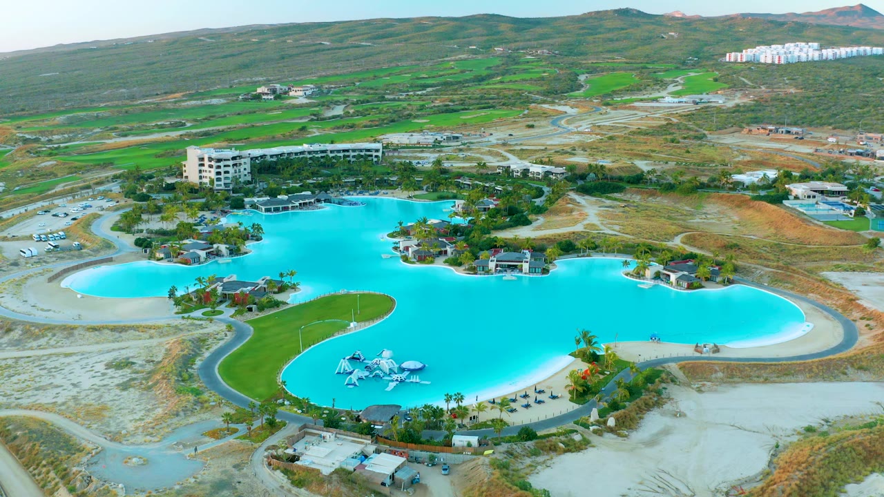 Drone view of the turquoise pool complex with a rounded beach-entry edge, slide structure, buildings, and hillside background.