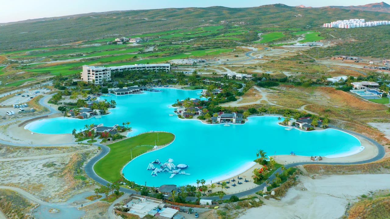 At 0:02, the aerial view keeps the blue water park pool broad in frame, with the slide cluster low in the composition, winding paths around the water, and green hills rising behind sandy development land.
