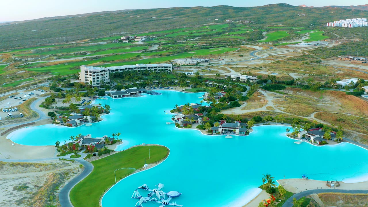 Aerial frame over Diamante Cabo San Lucas showing a bright turquoise lagoon, curved sandy edges, resort buildings, roads, green hills, and no visible people.