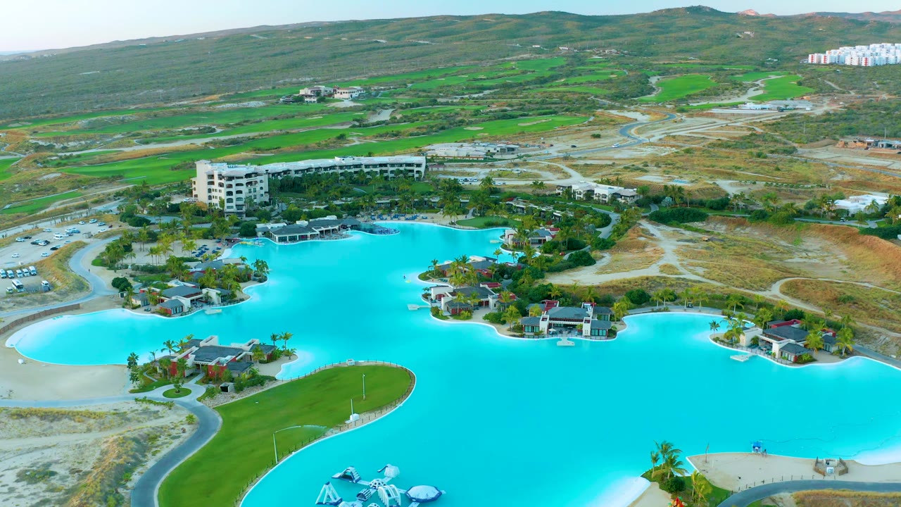 Aerial view of Diamante Cabo San Lucas with turquoise lagoon water, pale sandy curves, small shoreline buildings, a larger resort building, roads, hills, and no visible people.