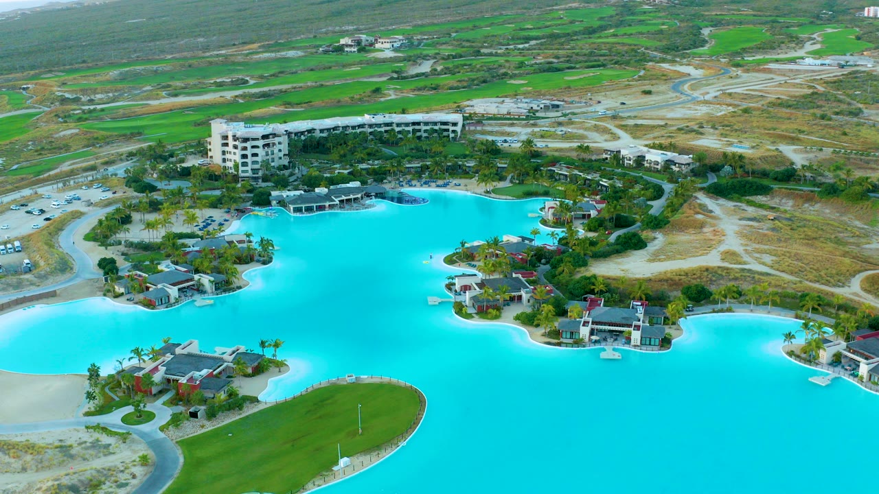 High aerial view over Diamante Cabo San Lucas showing a bright turquoise lagoon, small shoreline villas, a white resort building, green fairways, sandy roads, and no visible people.