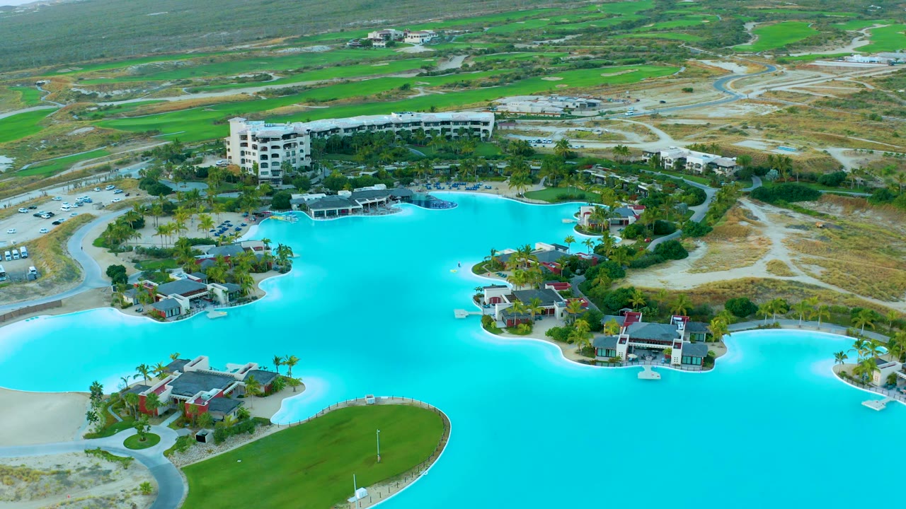 Drone aerial over Diamante Cabo San Lucas showing the turquoise lagoon across the lower frame, villas on small peninsulas, a multi-story resort block, golf course patches, roads, and no visible people.