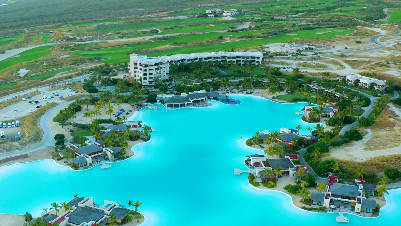 High aerial view over Diamante Cabo San Lucas with a turquoise lagoon in the foreground, resort buildings and a parking lot beyond it, green fields in the distance, and no visible people.