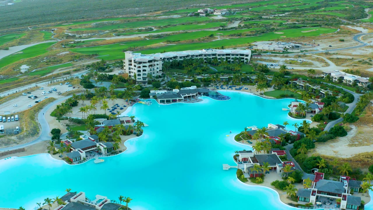 High oblique aerial of Diamante Cabo San Lucas with the lagoon spreading across the foreground, a large resort building near the center, parking and access roads to the side, and no visible people.