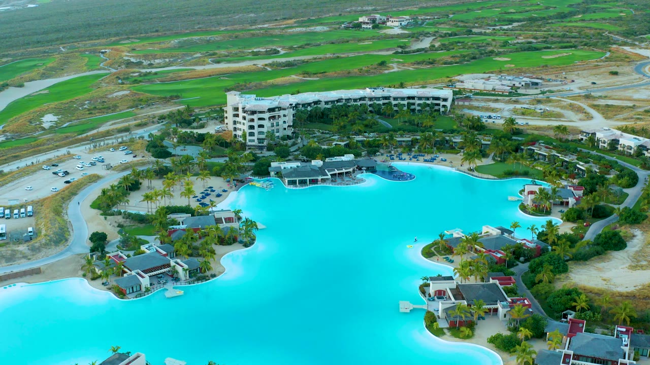 Aerial view of Diamante Cabo San Lucas where the turquoise lagoon occupies the lower frame, resort structures and palms sit along the water, roads and green fields fill the background, and no visible people appear.