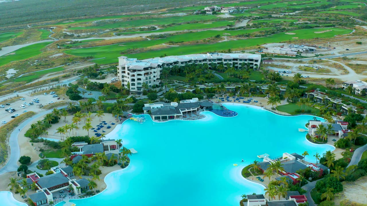 Aerial view at 0:01 over Diamante Cabo San Lucas with the bright pool centered lower frame, palm-lined paths, a white resort building, parked cars, roads, and no visible people.