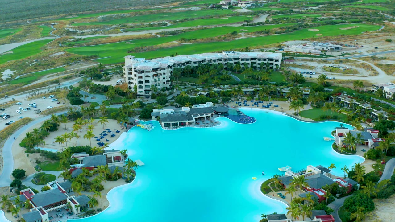 Aerial frame over Diamante Cabo San Lucas showing the pool occupying the lower half, small resort structures at the edge, parking and vehicles to the left, green landscape beyond, and no visible people.