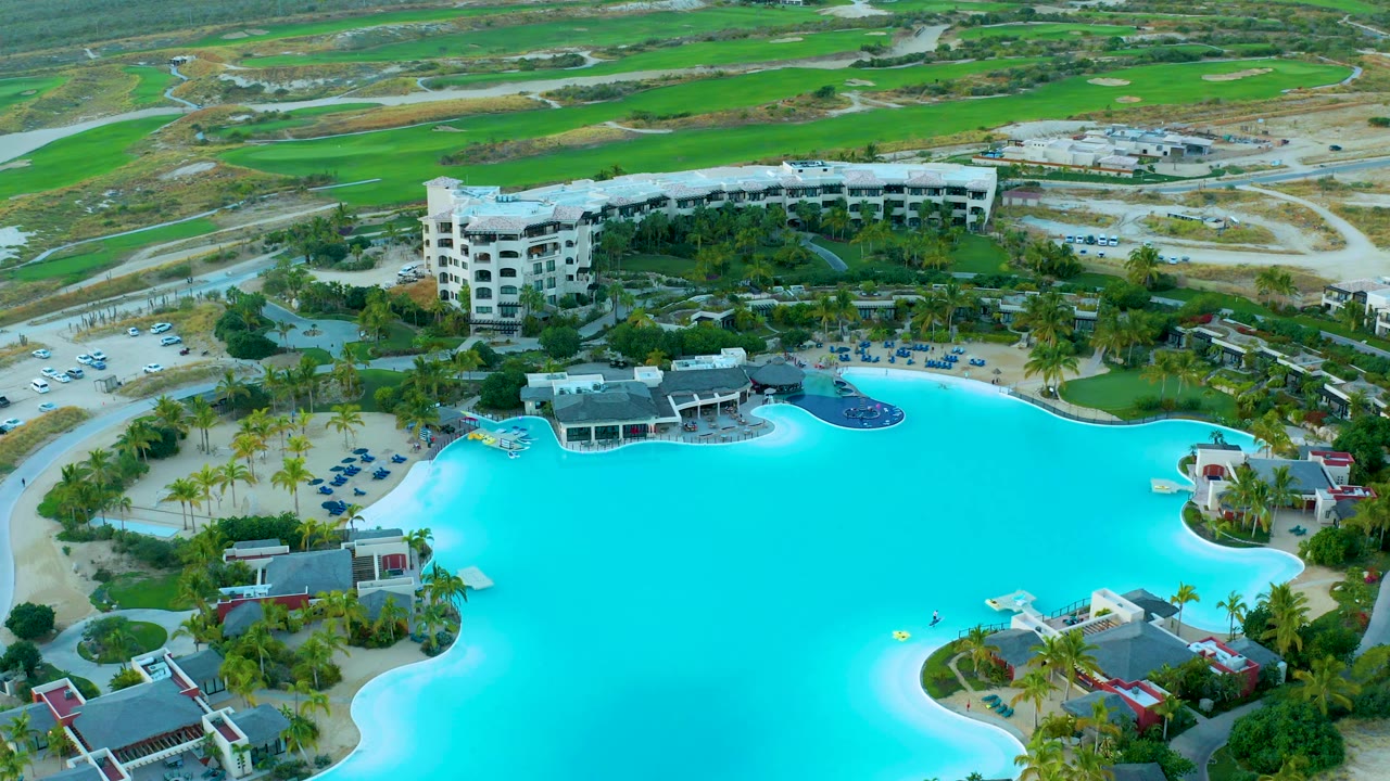 Aerial view at 0:03 in Diamante Cabo San Lucas with the curved pool edge, palm clusters, poolside buildings, a white resort block, cars in the lot, open roads, and no visible people.