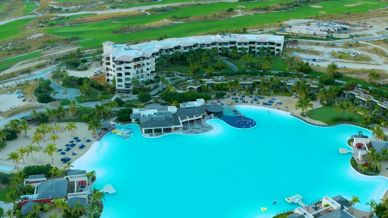 Aerial frame at Diamante Cabo San Lucas showing a large turquoise resort pool, a yellow float near the water, palm trees, lounge chairs, buildings, roads, and no visible people.