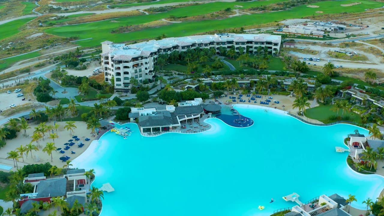 Aerial view in Diamante Cabo San Lucas with the pool filling the lower frame, the yellow float on the water, palm-lined paths, a multi-story building, parked vehicles, and no visible people.