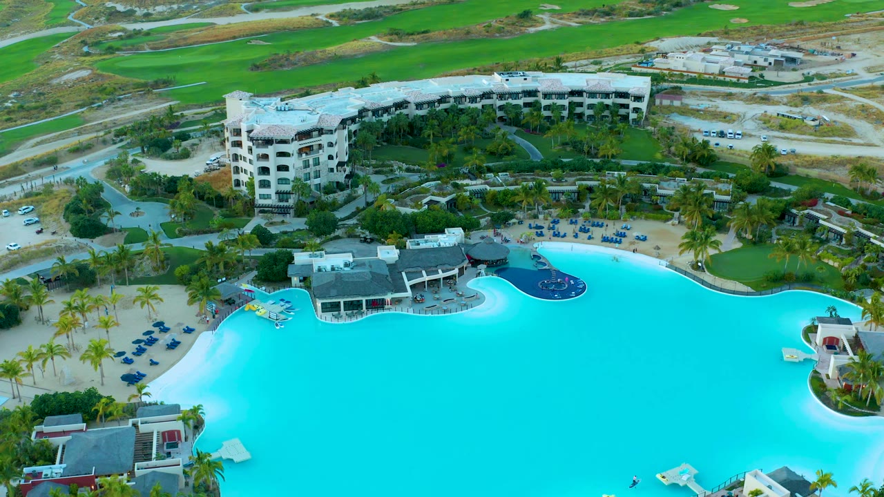 Aerial frame from Diamante Cabo San Lucas showing the curved turquoise pool edge, a small yellow float, dark blue pool section, palms, lounge chairs, resort buildings, roads, and no visible people.