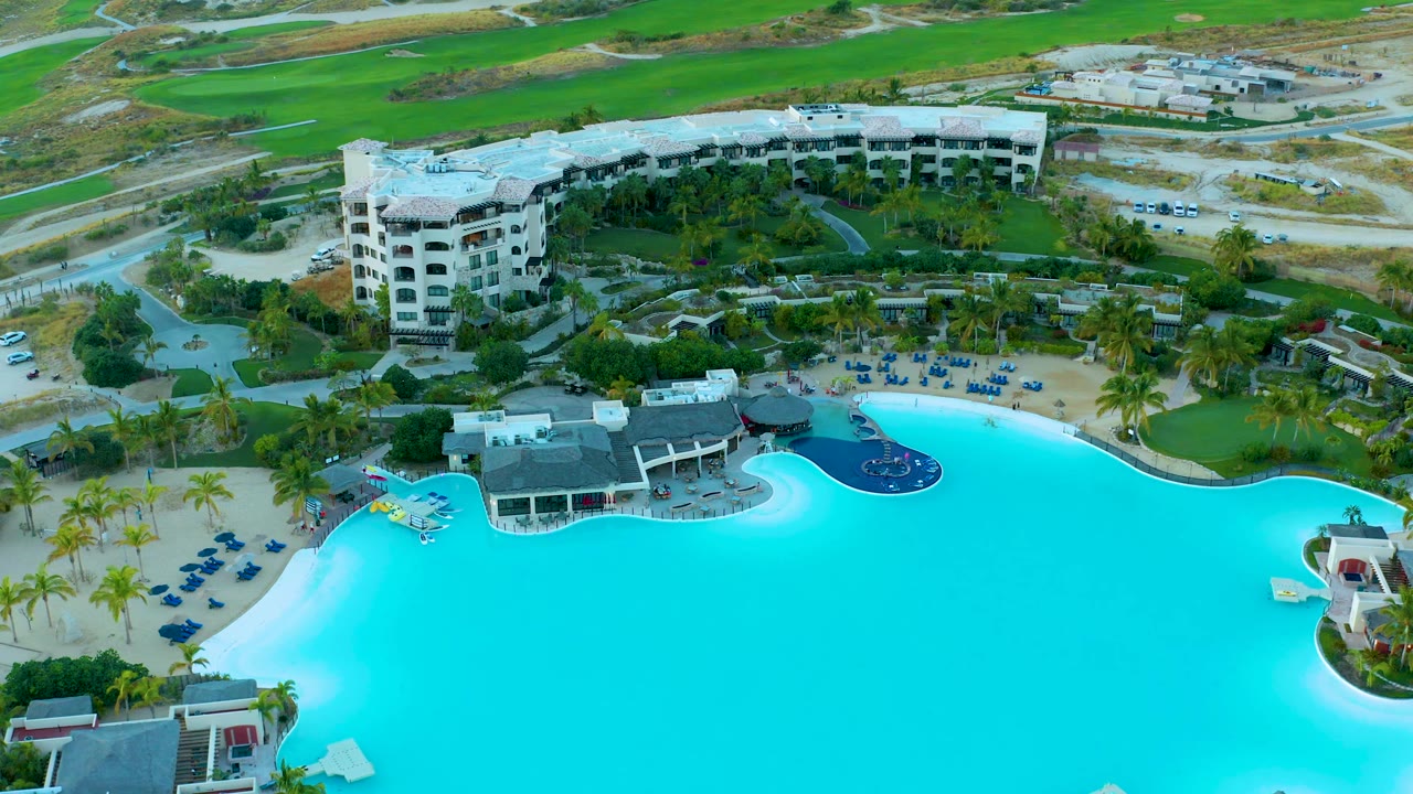 Drone view of resort pool, lounge chairs, palm trees, and a multi-story building.