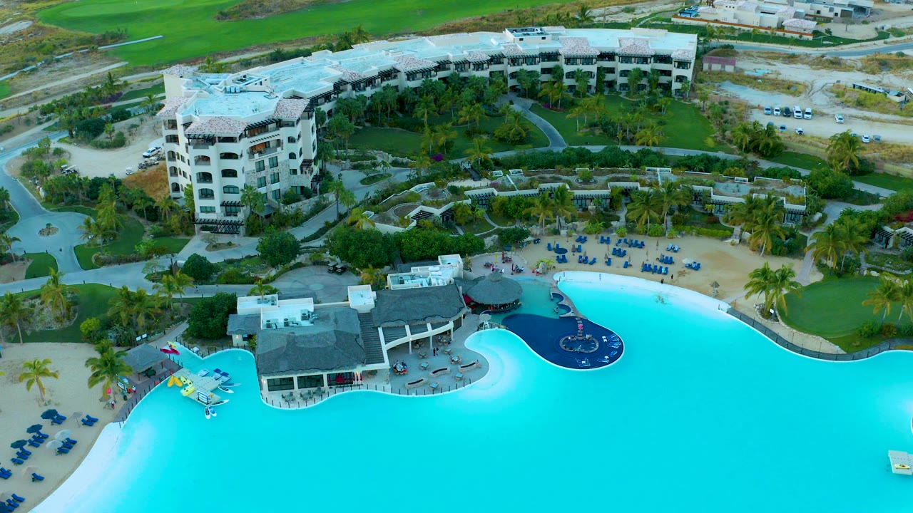 Aerial frame over Diamante Cabo San Lucas showing a large turquoise pool in the foreground, blue lounge chairs on sand, palms, landscaped paths, and white resort buildings with no recognizable people.