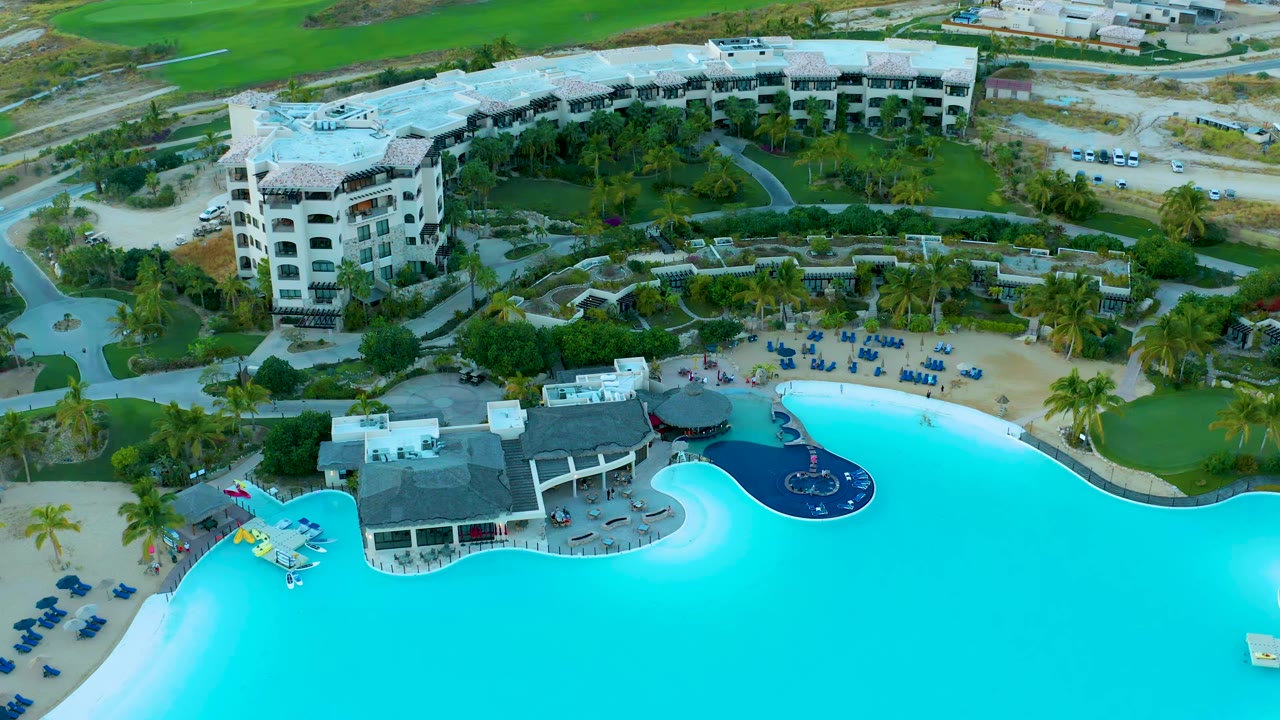 Aerial view at Diamante Cabo San Lucas with the pool filling the lower frame, a dark circular water feature near the deck, rows of lounge chairs on sand, palms, and resort buildings beyond.
