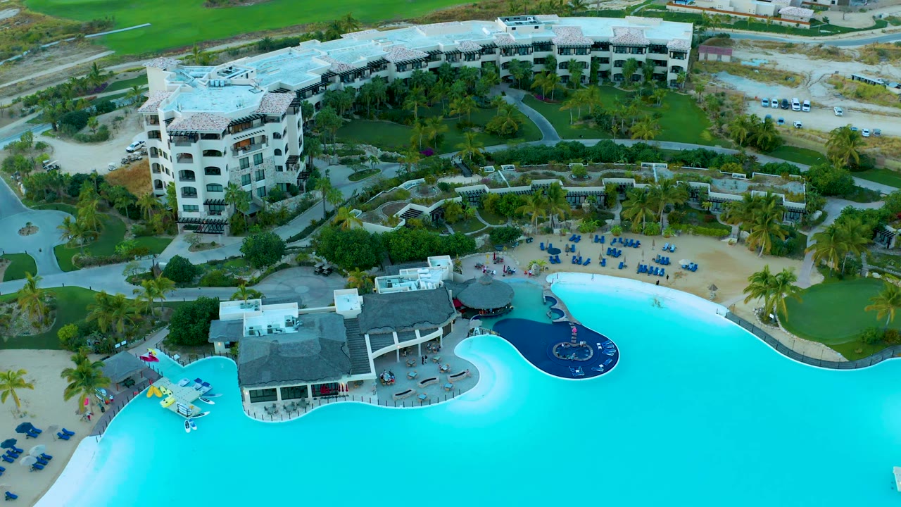 Aerial frame from the Diamante Cabo San Lucas resort area showing bright blue pool water across the bottom, a sandy chair area, palms, curved paths, and white multi-story buildings in the background.