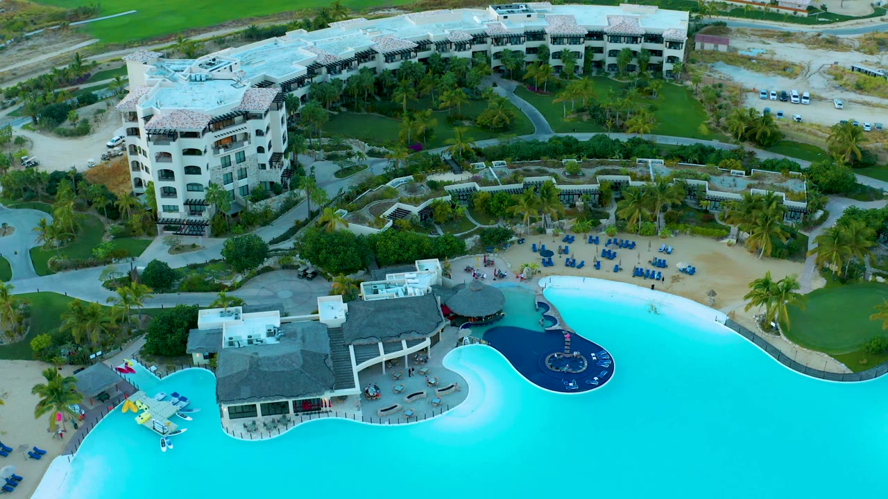 Aerial resort pool beside a sandy lounge area with palms and white buildings.
