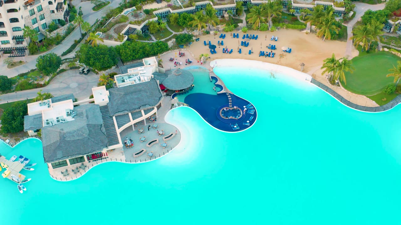 Aerial view in Diamante Cabo San Lucas with the bright pool across the lower frame, a curved dark-blue pool section near the center, beach lounge chairs above it, resort buildings, greenery, and no visible people.
