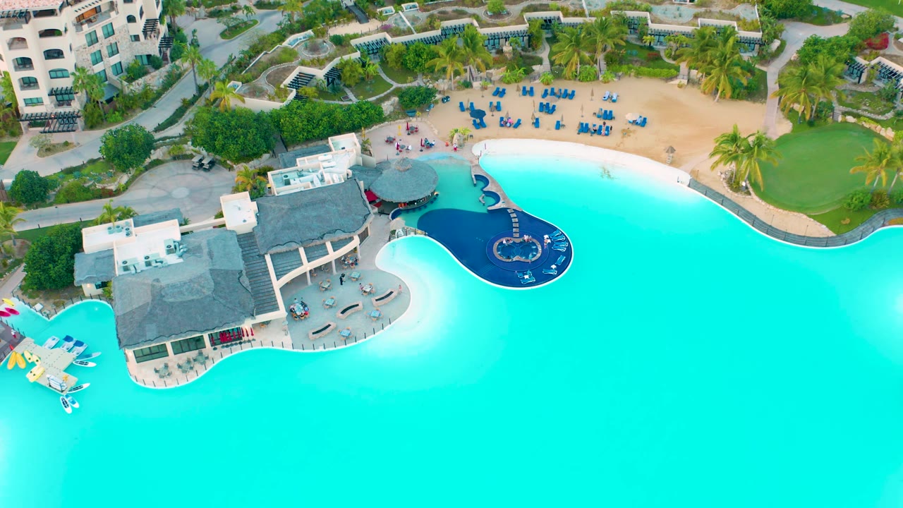High drone frame over Diamante Cabo San Lucas showing more of the pool's open turquoise water on the right, the thatched poolside buildings near center, sandy lounge area above, planted grounds, and no visible people.