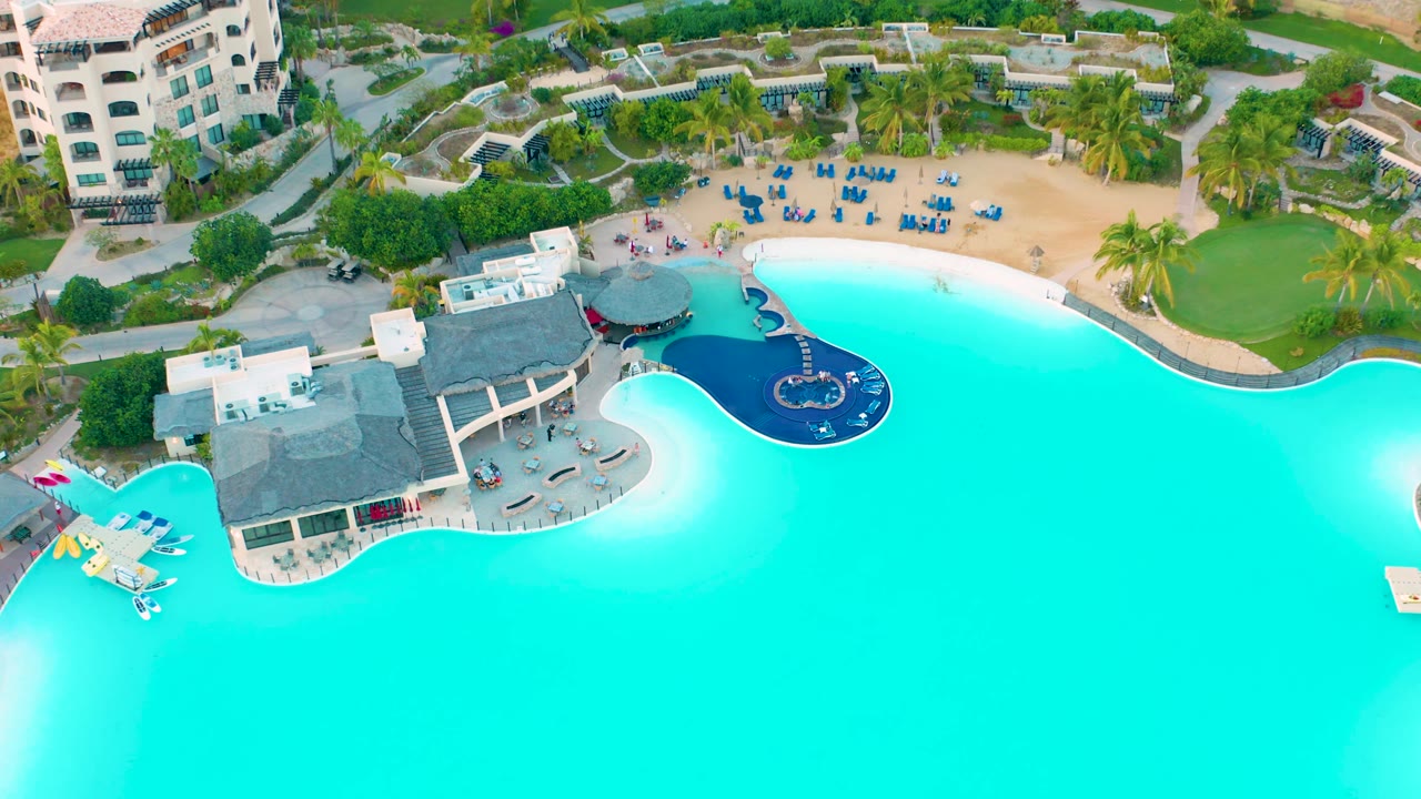 Drone frame in Diamante Cabo San Lucas with the pool sweeping across the lower half, a cluster of thatched-roof structures at left center, a sandy beach with blue chairs above, resort buildings and planted paths behind, and no visible people.