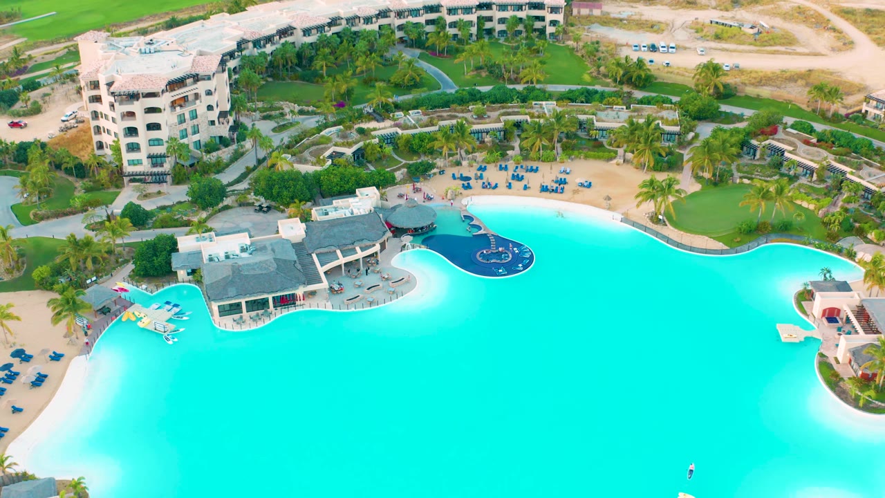 Second aerial frame at Diamante Cabo San Lucas with the pool still centered, a poolside building at left, lounge chairs above the water, a road and green field in the background, and no visible people.