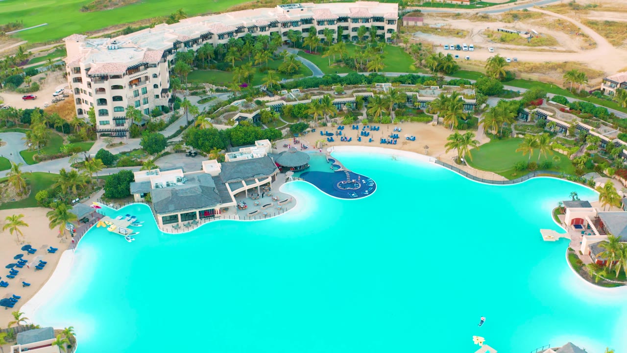 Aerial frame over the Diamante Cabo San Lucas resort pool, with deep blue pool details near the upper edge, turquoise water below, landscaped grounds, roads, open dirt areas, and no visible people.