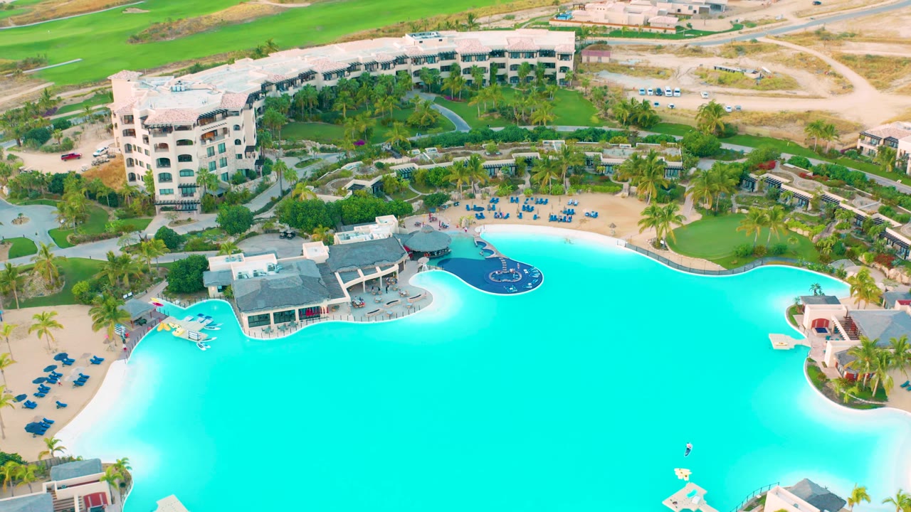 Aerial view of resort pool, poolside structure, palms, chairs, and road.
