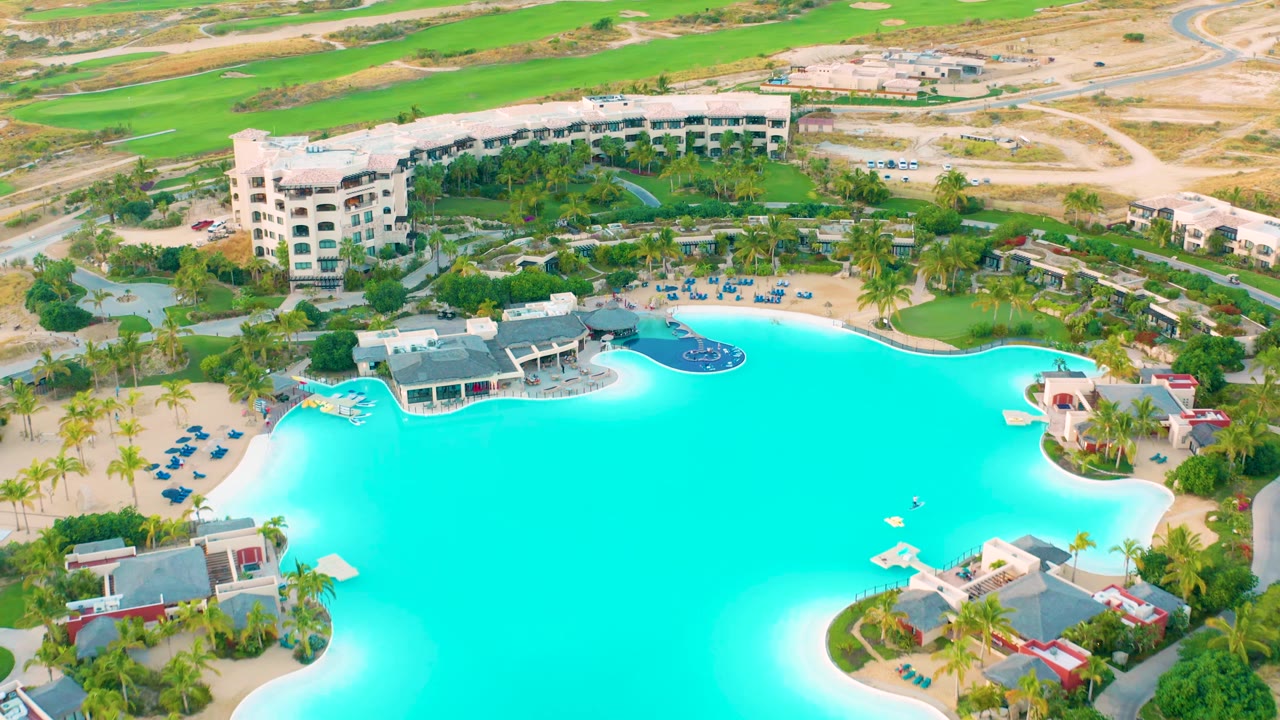Aerial view in Diamante Cabo San Lucas with the turquoise pool spanning the center, a curved shoreline-style edge, low buildings, palms, paths, and no visible people.