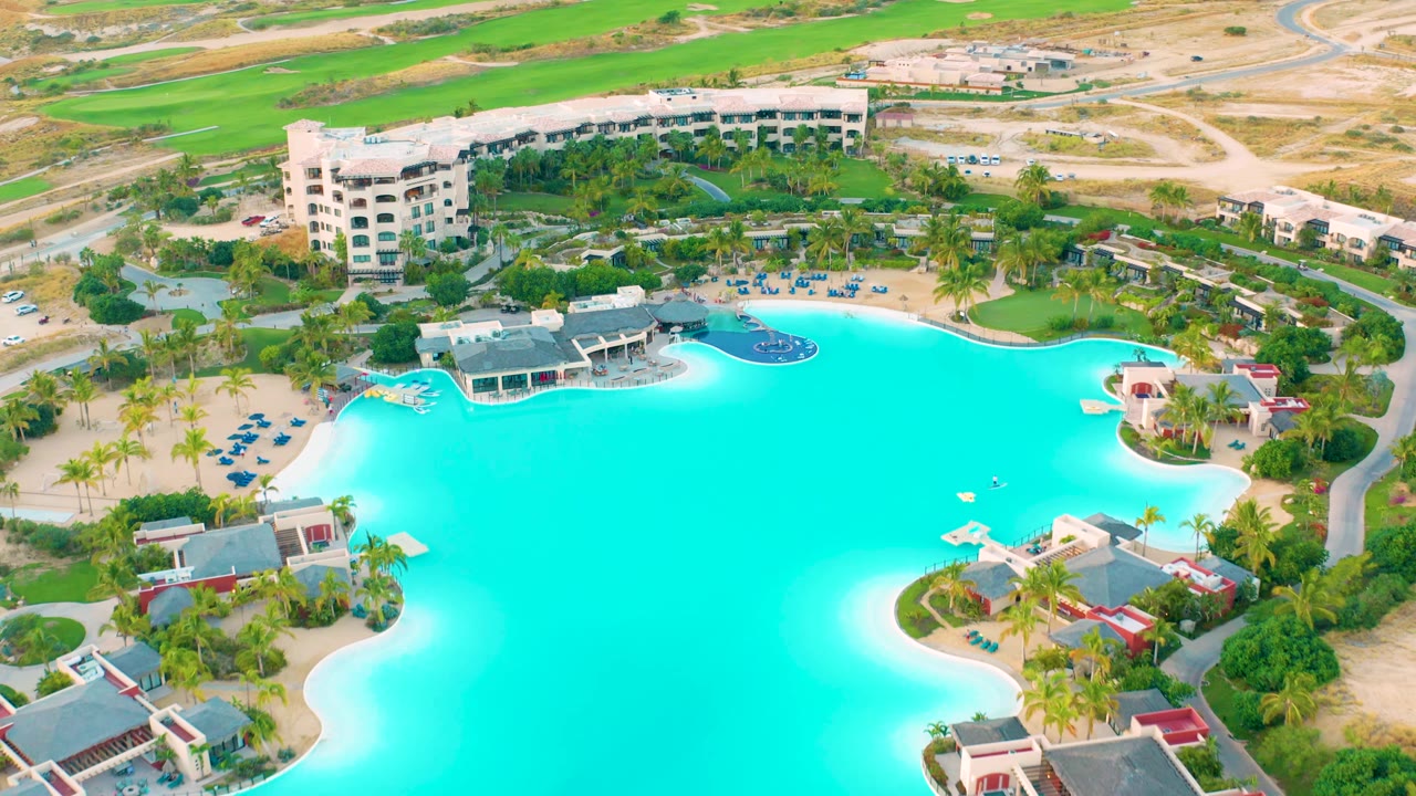 Aerial frame over Diamante Cabo San Lucas showing the turquoise pool, a dark circular pool feature near the upper edge, clustered buildings, palms, and no visible people.