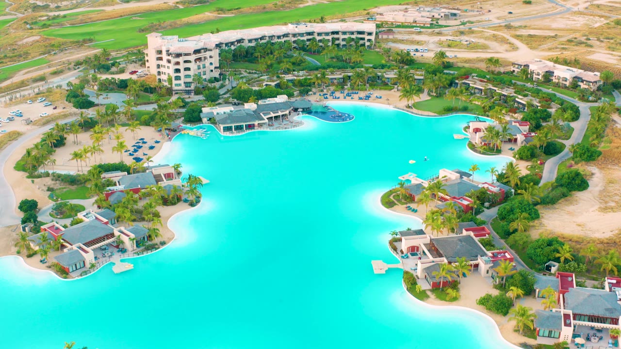 Aerial view of a turquoise lagoon at Diamante Cabo San Lucas, with sandy shoreline, low resort buildings, palm trees, roads, dry terrain, and no visible people.
