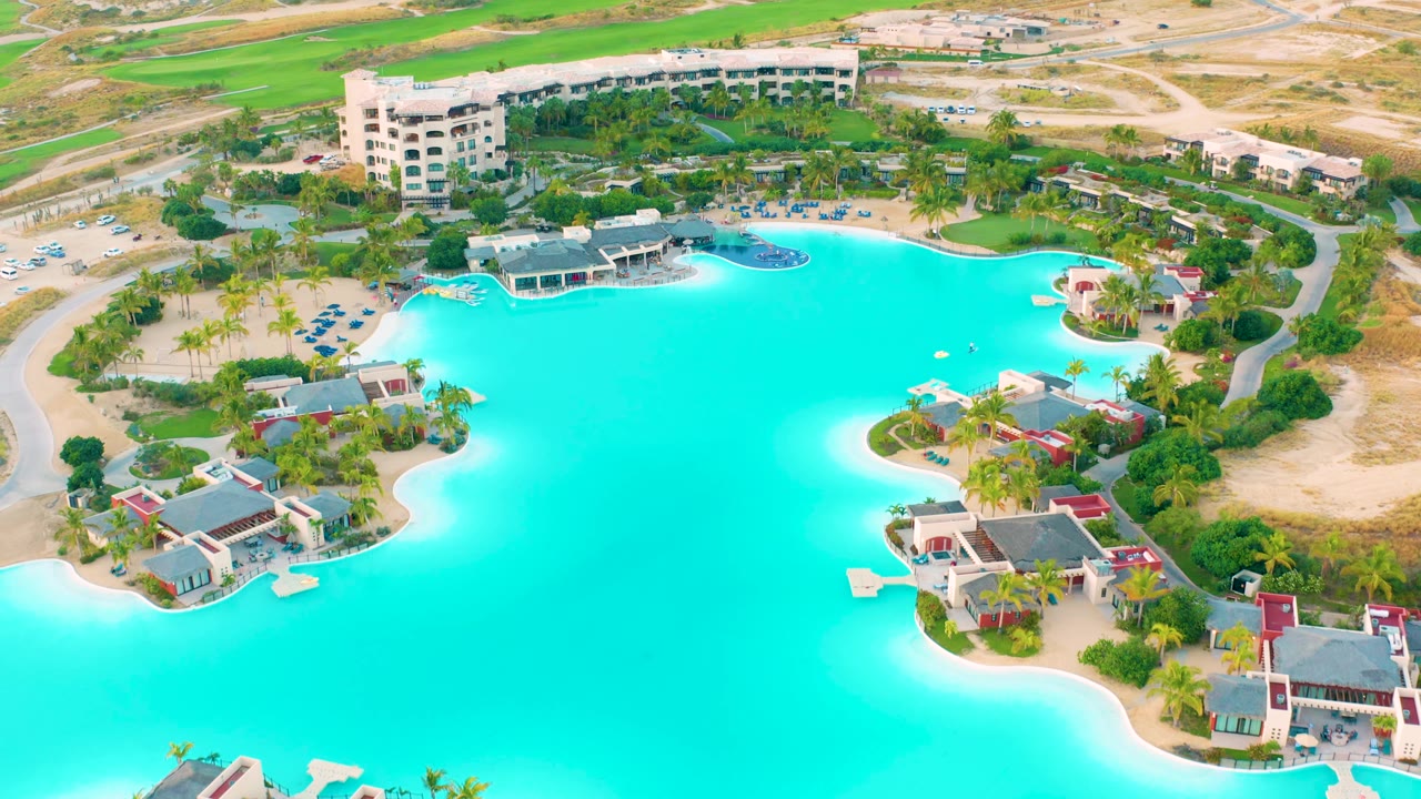 Aerial view in Diamante Cabo San Lucas with the turquoise lagoon stretching across the frame, sandy banks, clustered resort buildings at lower right, greenery, and no visible people.