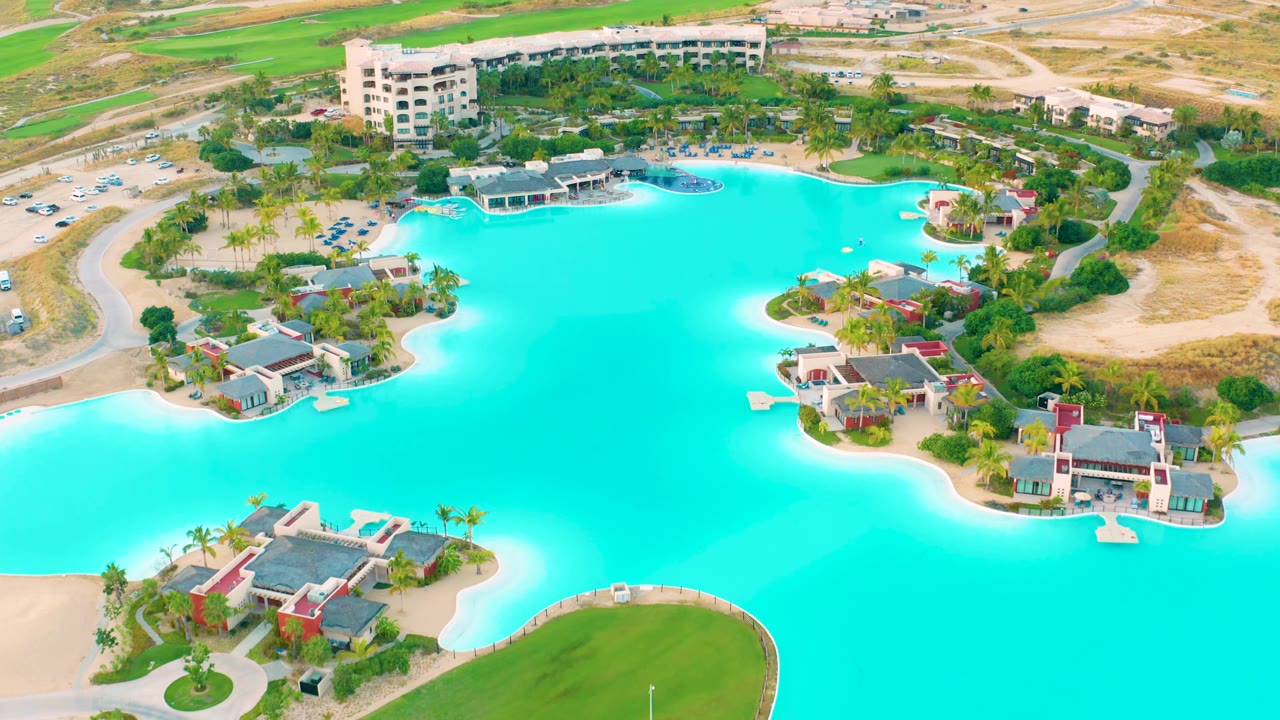 Early aerial frame over Diamante Cabo San Lucas with the turquoise lake filling most of the view, villas on small shorelines, roads and parking above, and no visible people.