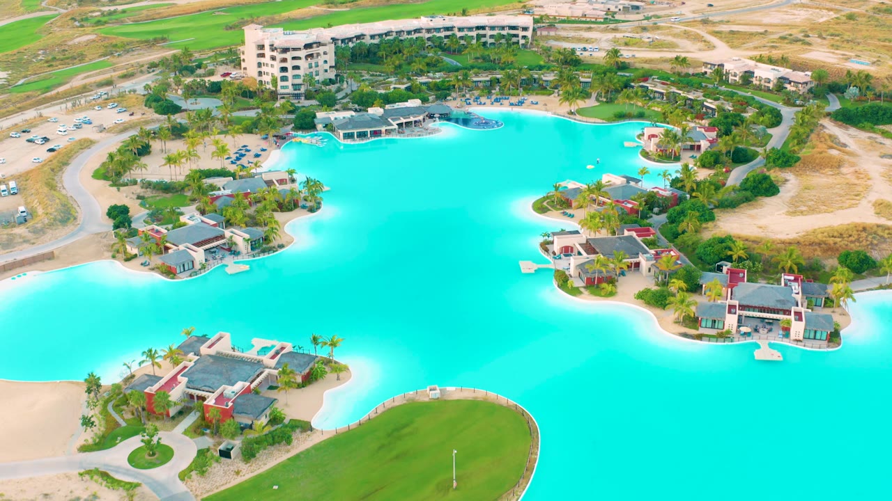 Aerial frame in Diamante Cabo San Lucas showing saturated turquoise water, a rounded green lawn in the foreground, villas along the shore, distant buildings, and no visible people.