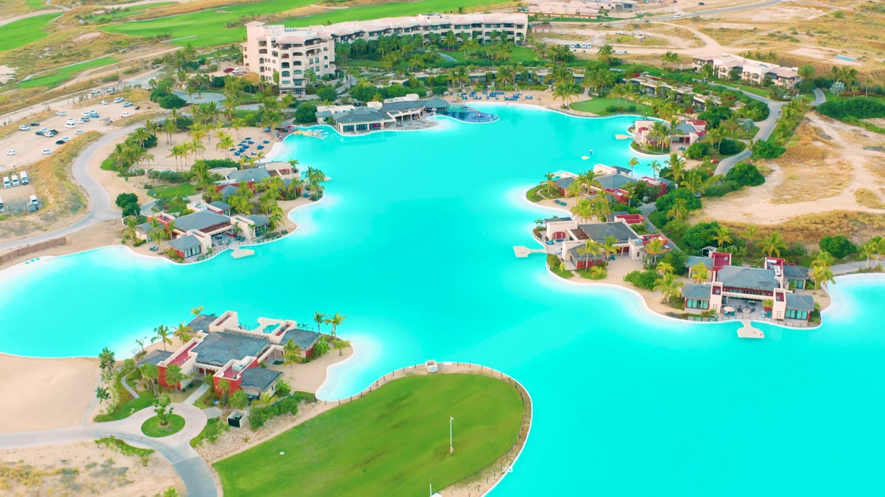 Aerial lake view with turquoise water, homes, road curves, parking, and dry terrain.