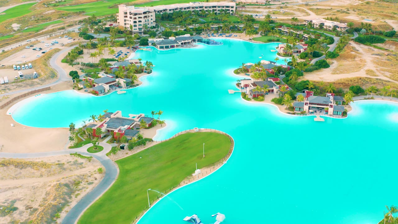 Opening aerial frame over Diamante Cabo San Lucas shows turquoise lagoon water, a green lawn peninsula, curved road, sandy ground, resort buildings, palm landscaping, and no visible people.